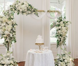 Elegant white floral wedding arch with drapes and three-tiered cake in a bright room—ideal for Louisiana weddings.