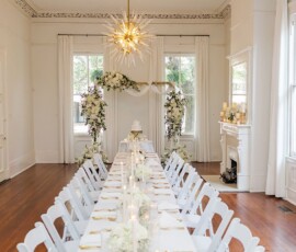 Elegant wedding table in a bright white room with wood floors, floral decor, and cake beneath arches; gold chandelier overhead.