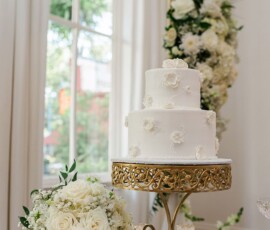 Elegant white two-tier cake with floral decor on a gold stand, surrounded by roses and candles at a New Orleans wedding.