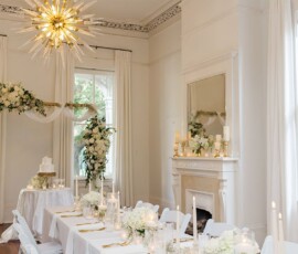 White wedding room with decorated dining table, white flowers, candles, cake, gold chandelier, and floral arch by large windows.