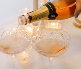 Hand pours sparkling wine into two coupe glasses on a white table with glowing candles, creating an elegant wedding vibe.