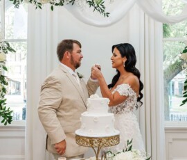 Bride and groom feed each other cake amid white flowers, drapery, and large windows at their elegant Louisiana wedding.