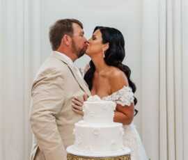 Bride and groom kiss by a white wedding cake on gold stand, surrounded by white flowers, candles, and elegant drapery.