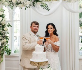 Bride and groom toast by a white cake amid elegant white flowers and drapes—perfect inspiration for Louisiana weddings.