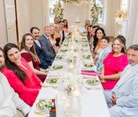 Formally dressed guests smile and pose at a long table with salads, candles, and white flowers at an indoor wedding reception.