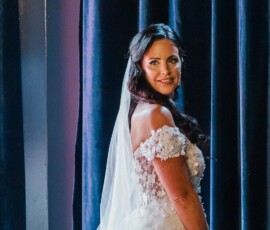 Woman in a white floral wedding dress and veil smiles at the camera before deep blue curtains, exuding New Orleans elegance.