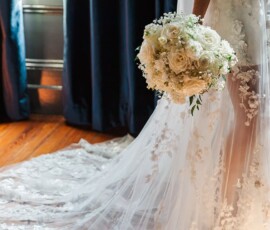 Bride in lace gown stands on wooden floor with white rose bouquet, sunlight on her train before New Orleans wedding.