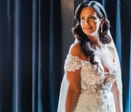 Bride with long dark hair in off-shoulder lace gown smiles by dark curtains, capturing New Orleans wedding inspiration.
