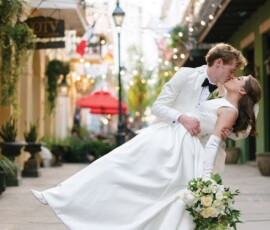 Groom in tuxedo dips and kisses bride in white dress on a decorated street; she holds a classic white bouquet.