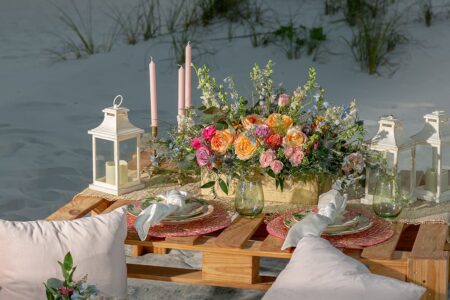 Wooden picnic table on sand with floral centerpiece, glassware, pink napkins, lanterns, and pillows—ideal for Louisiana weddings.