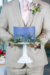 Person in beige suit holds purple cake with gold edges and thistle on white stand—elegant for a Louisiana wedding.