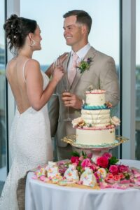Bride and groom smile by a table with wedding cake and treats, holding champagne flutes at their reception near large windows.