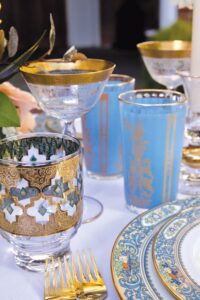 Luxurious table with gold-trimmed glasses, blue and gold plates, gold flatware, and a floral centerpiece—ideal for weddings.