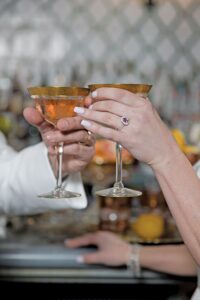 Hands with diamond ring and gold band clink gold-rimmed cocktails at a Louisiana wedding, blurred bar in background.