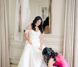 Bride in white smiles as woman in pink helps with shoes in elegant room before Indian wedding at Ritz-Carlton New Orleans.