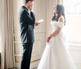 Bride in white gown smiles at emotional groom wiping his eye by a window at their Ritz-Carlton New Orleans wedding.