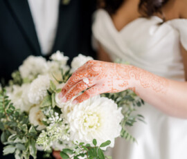 A bride’s henna-adorned hand, wearing her engagement ring, gently rests on a bouquet of white dahlias and greenery. Part of her off-the-shoulder white gown and the groom’s black tuxedo are softly visible in the background.