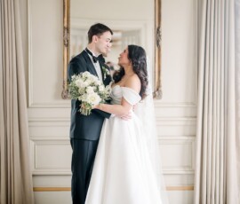 Bride and groom stand smiling indoors by a large ornate mirror and cream curtains; bride holds white bouquet.