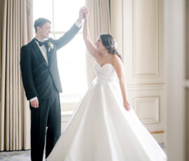 Bride in white gown and groom in black tuxedo dance by a window at their elegant, multicultural New Orleans wedding.