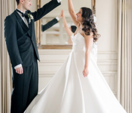 A bride and groom dance joyfully in an elegant room, with the bride twirling in her strapless white gown and veil, and the groom in a black tuxedo holding her hand. Behind them, a large ornate mirror and chandelier add to the romantic, formal atmosphere.
