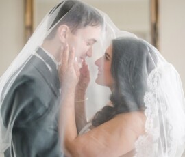 Bride and groom smile under a sheer veil, sharing a romantic moment with soft light and a mirror at their New Orleans wedding.