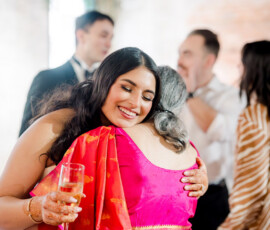 A smiling woman in a bright red and gold sari hugs an older woman during a celebration, holding a champagne glass in one hand. The moment is warm and joyful, with other guests chatting in the softly blurred background.