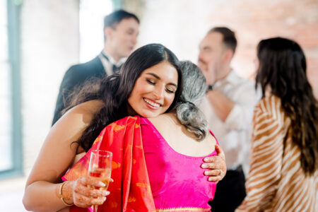 Woman in bright pink saree hugs another, smiling and holding a drink at a joyful Louisiana wedding; guests chat behind.
