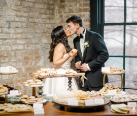 A bride and groom share a sweet kiss in front of a dessert table filled with cookies and pastries. The bride in a white gown holds a lemon bar while the groom in a black tuxedo leans in, standing against a rustic brick wall and large window.