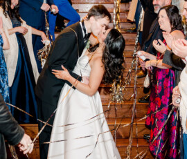 Bride and groom kiss at a staircase as guests with streamers cheer during their New Orleans Indian wedding.