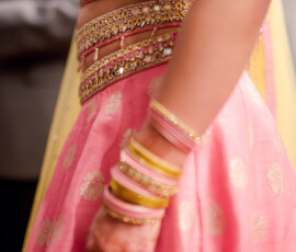 Close-up of a woman wearing a pink and gold embellished lehenga with mirrorwork details, henna on her hand, and stacked pink and gold bangles as she stands at an Indian pre-wedding celebration