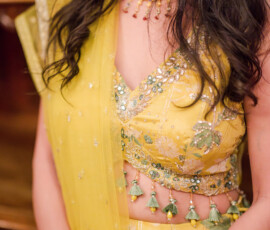 Close-up of a woman wearing a yellow embellished lehenga with floral embroidery, green tassel details, and matching green and gold bangles, paired with a gold necklace and soft waves in her hair