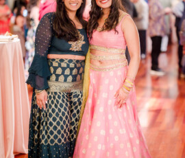 Two women in colorful Indian lehengas smile together at a lively New Orleans wedding, with guests and tables behind them.