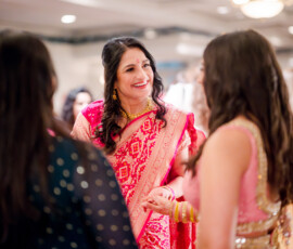 Woman in red and gold saree smiling at an indoor New Orleans Indian wedding, surrounded by guests in festive attire.
