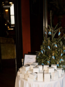 A decorated Christmas tree on a round table with white gift boxes and a “Please take one” sign, in warm dim lighting.