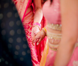 Two women in colorful sarees, one with henna, hold hands at a vibrant New Orleans Indian wedding in soft focus.