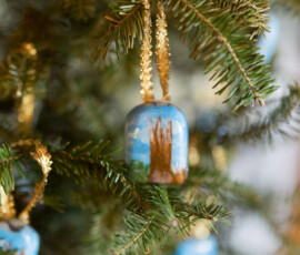 Blue hand-painted ornament with a brown building, hanging from a gold string on a Christmas tree, with blurred ornaments behind.