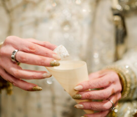 Person with glittery gold nails, rings, and ornate outfit holds a wooden cup and sugar cube at a multicultural New Orleans wedding.