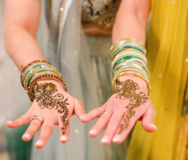 Two women extend their hands to show fresh mehndi designs on their palms, accented by colorful bangles. One wears a light blue lehenga and the other a yellow outfit, highlighting the intricate henna patterns.