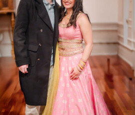 Couple smiling together at an Indian pre-wedding celebration; woman wearing a pink and gold lehenga with henna on her hands and traditional jewelry, standing beside her partner in a dark coat inside a warmly lit venue with polished wood floors.