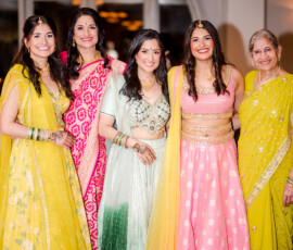 Group of women dressed in vibrant Indian attire at a pre-wedding celebration, wearing colorful lehengas and saris in yellow, pink, mint green, and red. They stand smiling together, showcasing traditional jewelry, henna designs, and elegant embroidery in a warm indoor venue.
