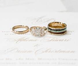 Close-up of three wedding rings displayed on top of an elegant ivory invitation with gold script lettering. The set includes a simple gold band, a three-stone princess-cut diamond engagement ring, and a gold men’s band with dark inlay and engraved date. Soft florals blur in the background.