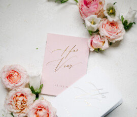 Pink and white flowers frame a pink Her Vows booklet and white box with gold writing in an elegant wedding flat lay.