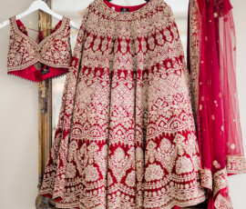 Red and gold embroidered bridal lehenga displayed on ornate hangers, including the heavily detailed skirt, matching embellished blouse, and sheer dupatta with gold trim, all hanging against an antique gold-framed mirror.