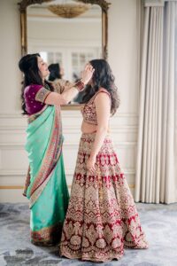 Two women in Indian attire prep for a Louisiana wedding, one adjusting the other’s hair, reflected in a large mirror.