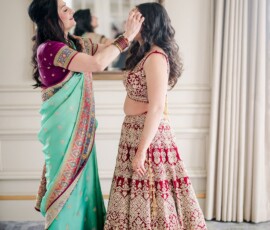 Two women in Indian attire prep for a Louisiana wedding, one adjusting the other’s hair, reflected in a large mirror.