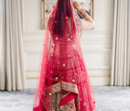 Bride seen from behind adjusting her red and gold embroidered dupatta, wearing a traditional lehenga with intricate beadwork and patterns. She stands in an elegant room with soft natural light, facing an ornate mirror and flowing curtains.