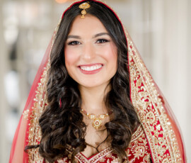 Bride smiling brightly while wearing a traditional red and gold embroidered lehenga with a matching dupatta draped over her head, featuring intricate beadwork and floral patterns. She has soft curls, elegant bridal jewelry, and a gold maang tikka, standing in soft indoor light.