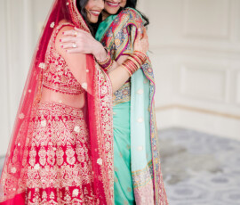 Bride in a red and gold embroidered lehenga warmly embraces a woman wearing a teal saree with colorful embroidered details. Both smile with eyes closed, sharing an emotional moment indoors before the ceremony.