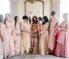 Bride in red and gold lehenga with six pink-clad bridesmaids smiling at her in a bright New Orleans wedding room.
