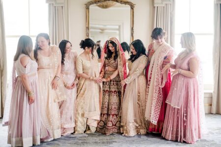 Bride in red and gold lehenga with six pink-clad bridesmaids smiling at her in a bright New Orleans wedding room.